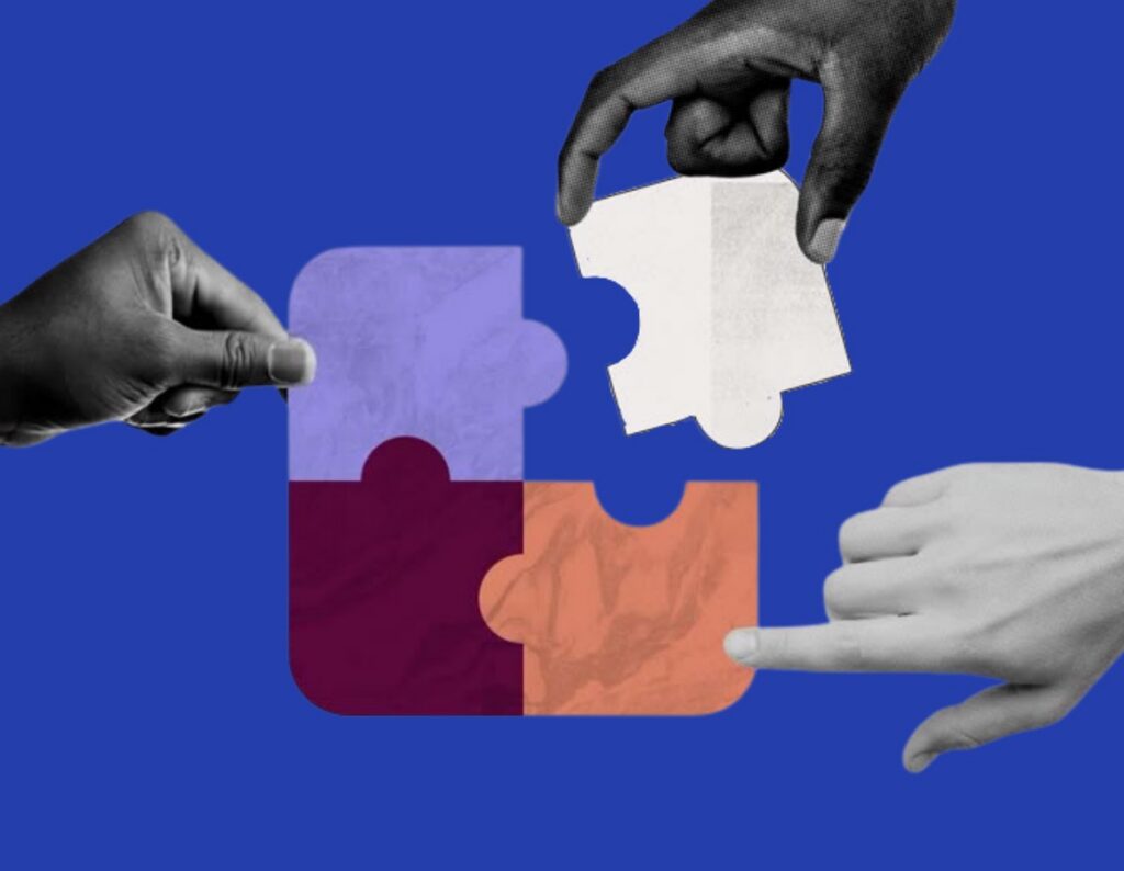 Four hands fitting coloured puzzle pieces together on the DAS royal blue, one piece still held apart from the rest. Lead image for the written piece on why DEI fails when it is treated as separate parts.